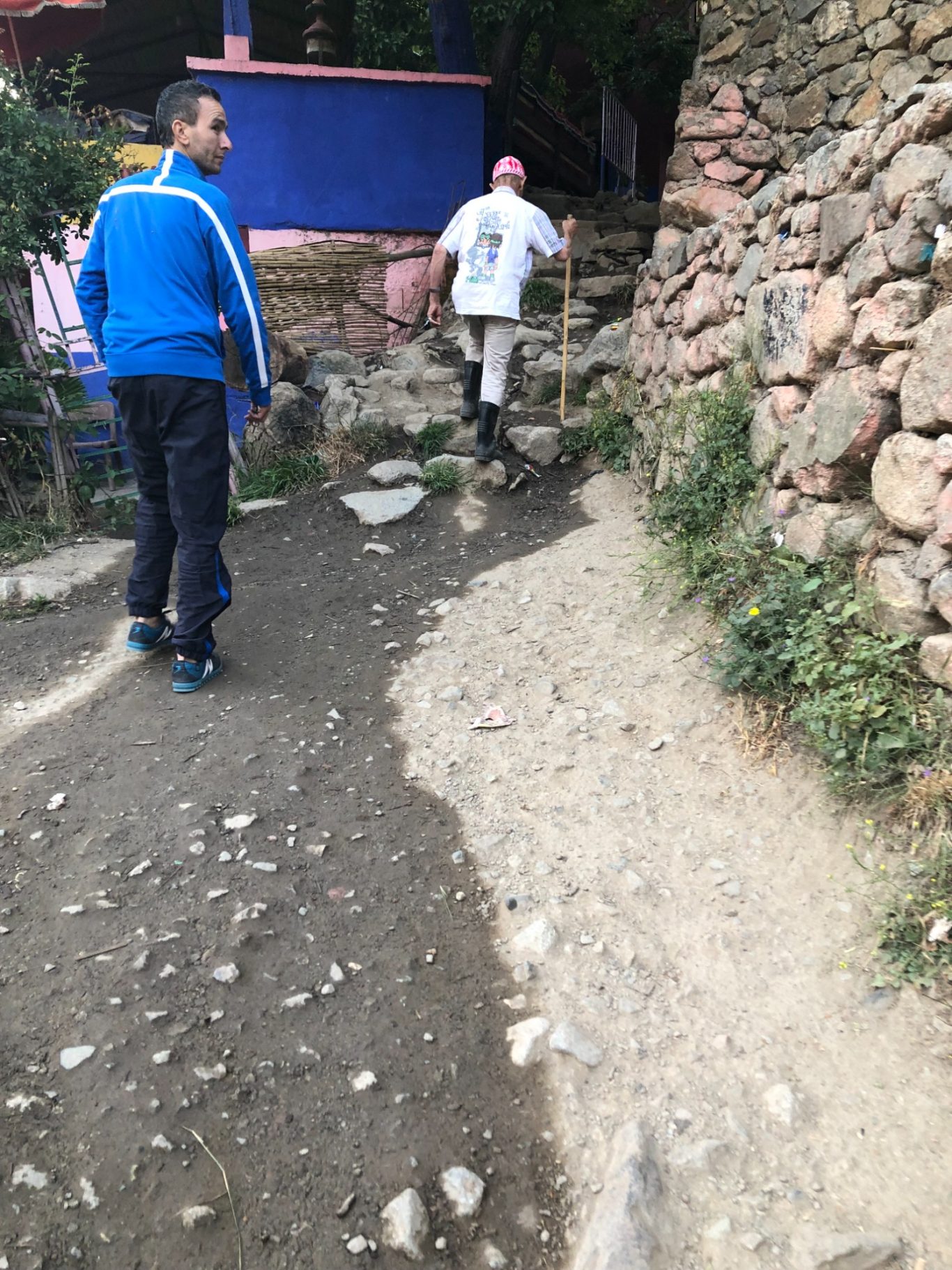 Two people walking along a rocky path with stone walls and a colourful building nearby.