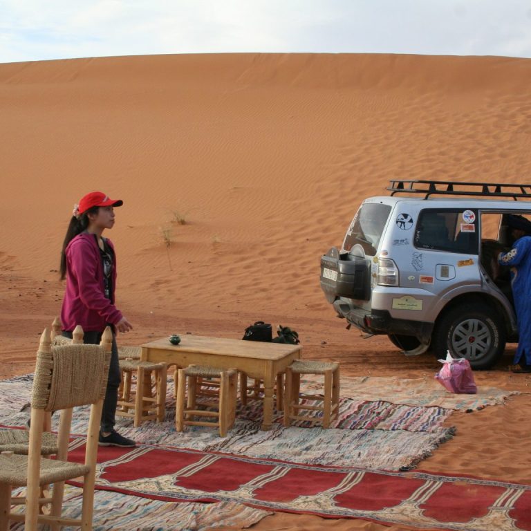 A desert scene with a woman by a 4x4 vehicle and a seated area on a rug.