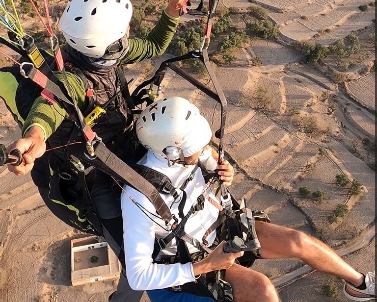 Two people paragliding above a landscape, one piloting and the other enjoying the view.