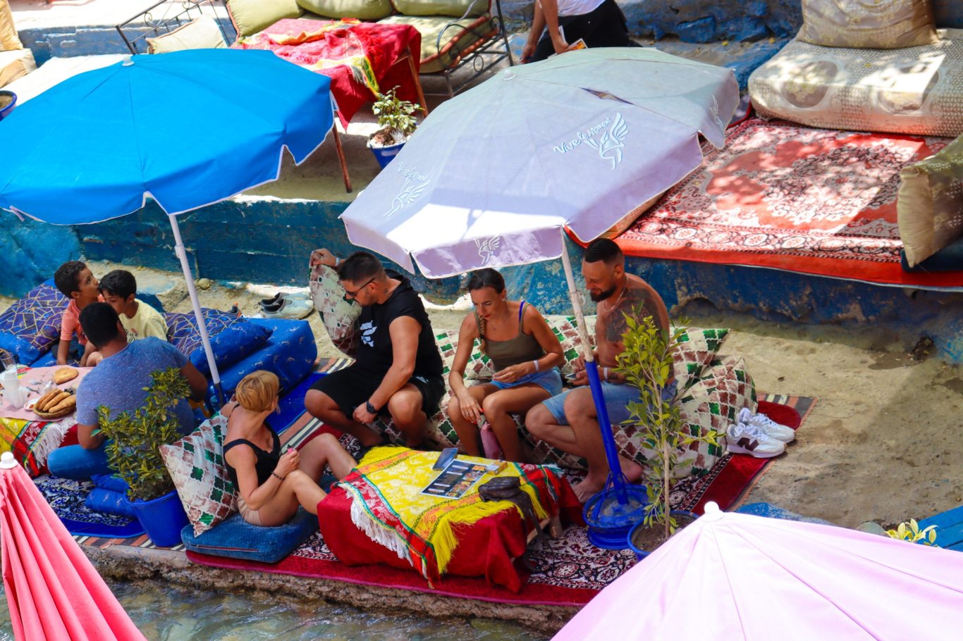 Group of people relaxing under umbrellas on a beach, surrounded by colourful cushions.
