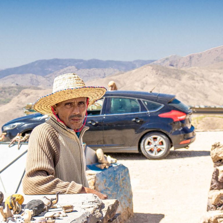 Man wearing a straw hat sits near a roadside, with mountains and a car in the background.