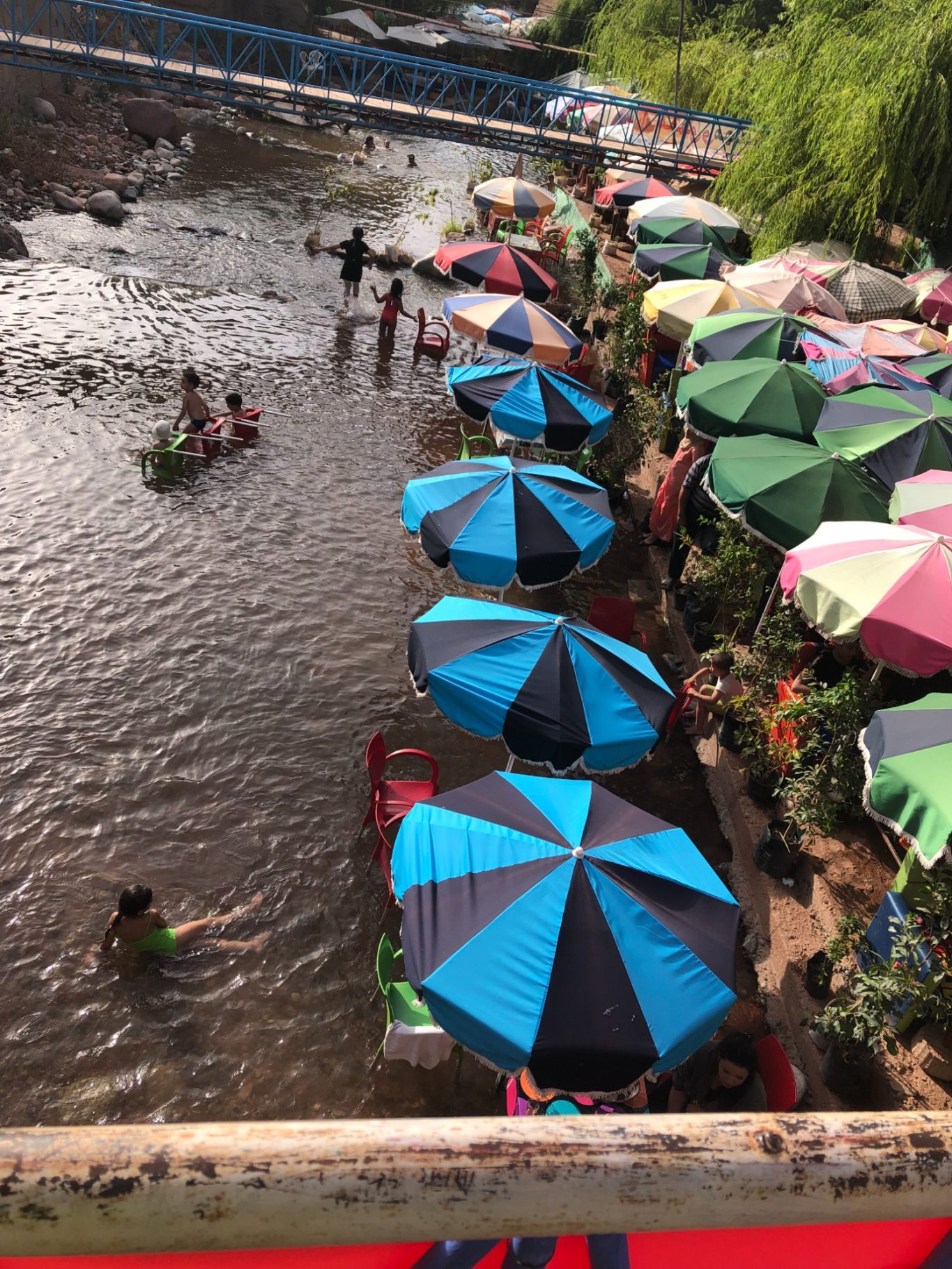 People enjoying a shallow river, with colourful umbrellas providing shade nearby.