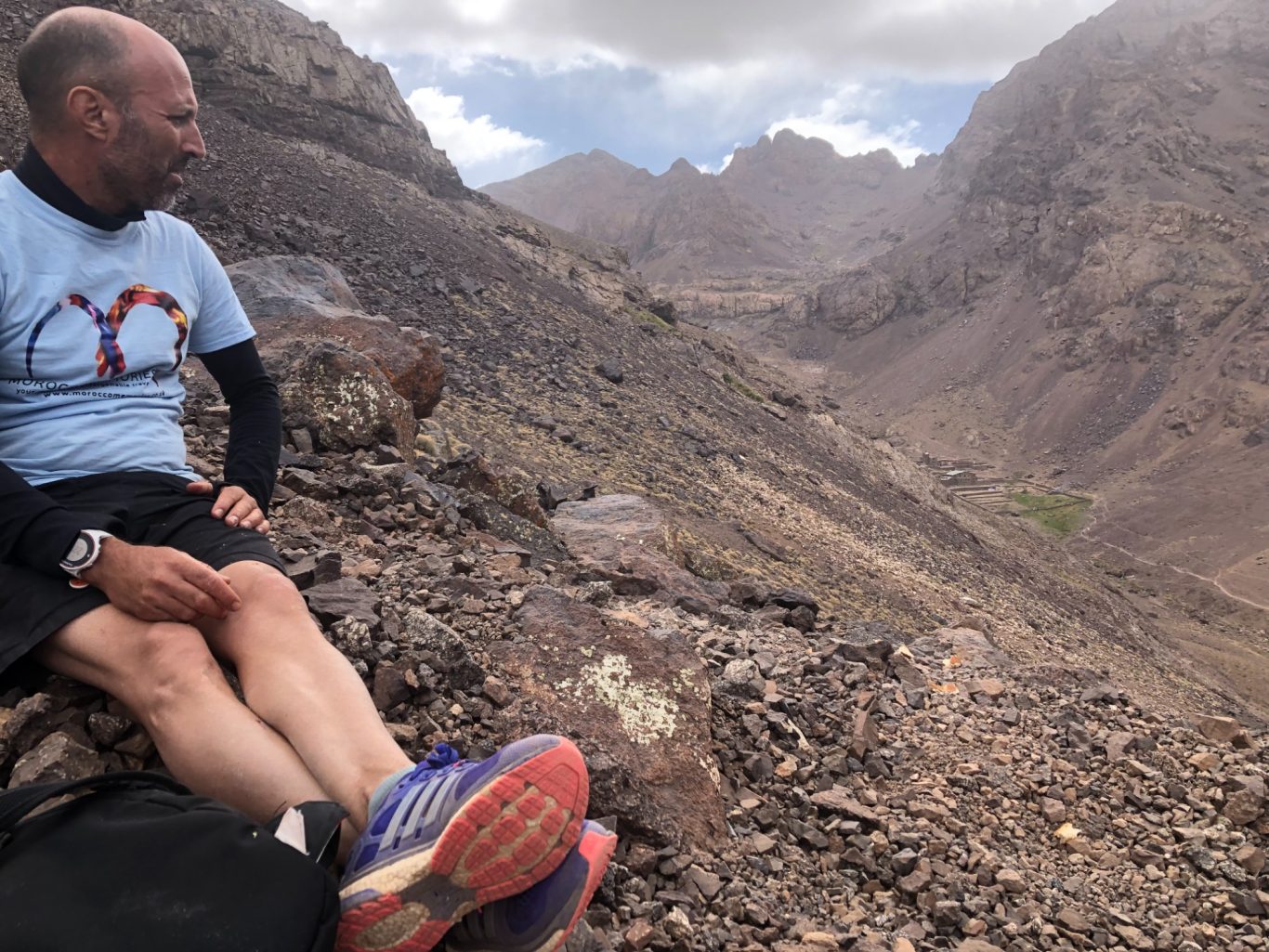 A man sits on rocky terrain, overlooking a mountainous valley under cloudy skies.