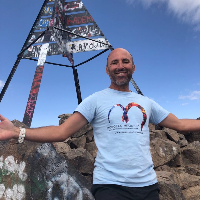 A man in a light blue T-shirt stands on rocks, smiling with arms outstretched near a painted structure.