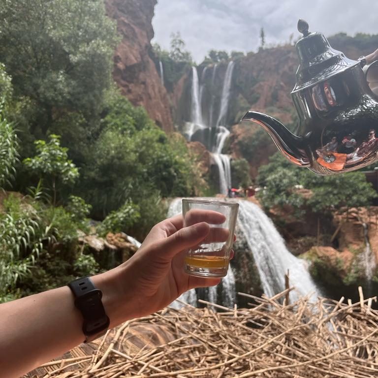A hand holding a glass of drink in front of waterfalls and a teapot in the background.