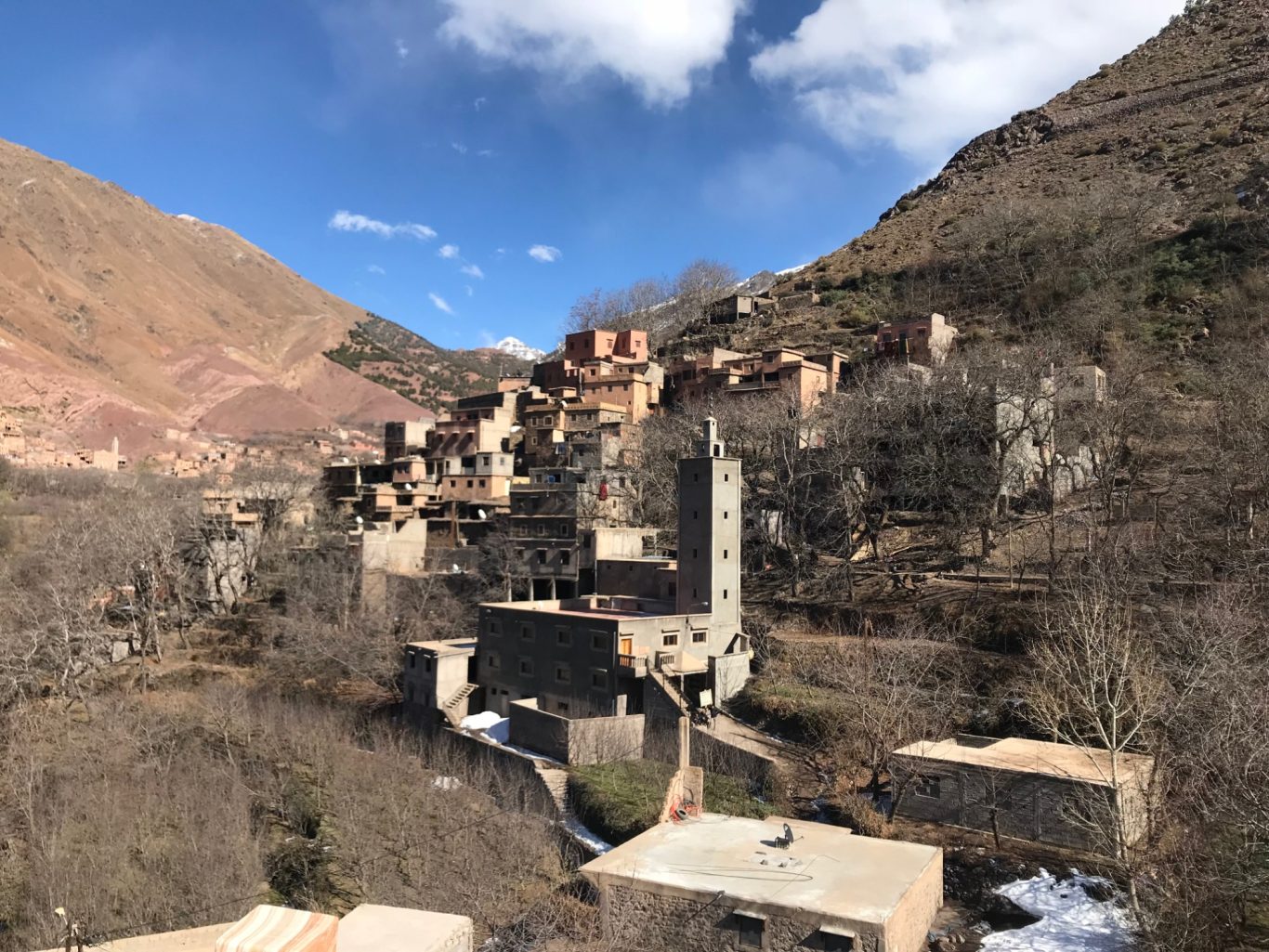 Mountain village with stone buildings, a church tower, and bare trees against a blue sky.