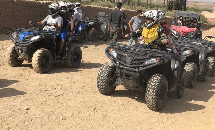 Several all-terrain vehicles lined up in a sandy outdoor location with riders preparing to depart.