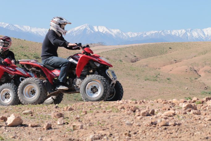 Two riders on quad bikes in a mountainous landscape with snow-capped peaks.