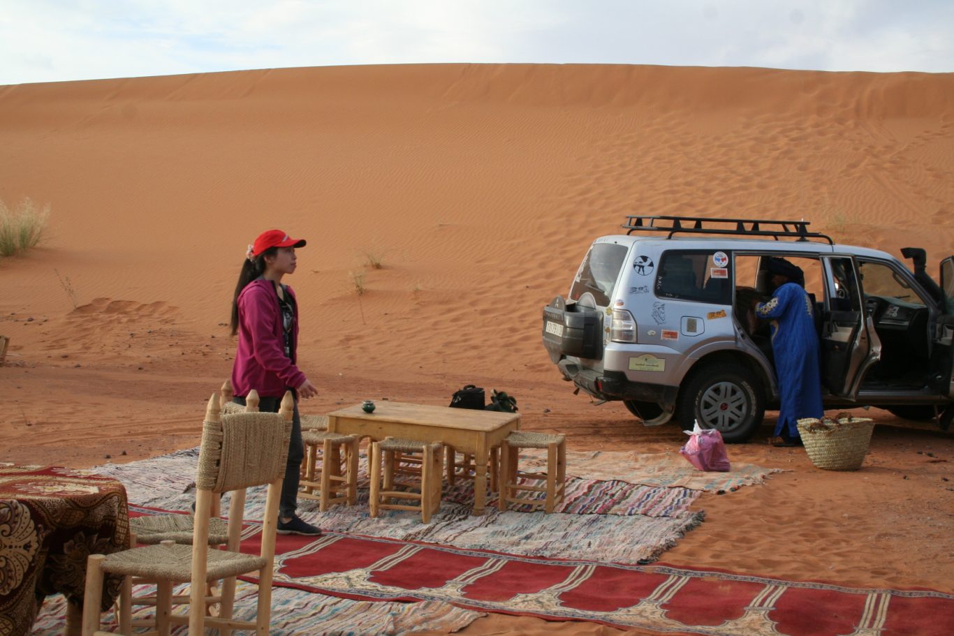A woman stands near a parked vehicle in a desert, surrounded by rugs and a table.