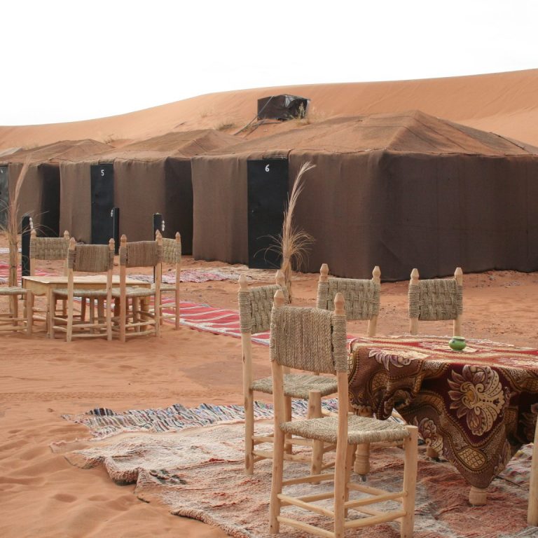 Traditional desert campsite with tents, wooden chairs, and a low table on sandy terrain.