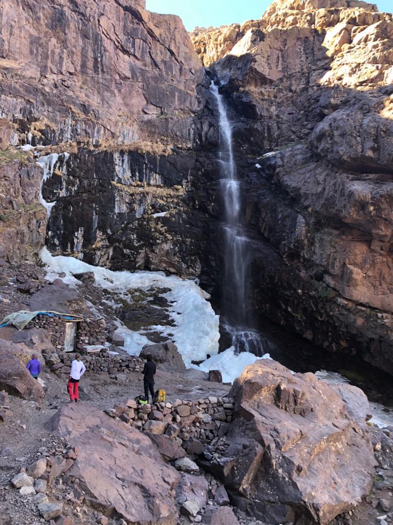 Waterfall cascading down rocky cliffs with viewers below. Snow patches visible.