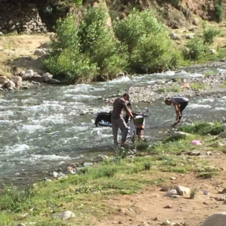 People wading in a shallow river, surrounded by greenery and rocky banks.