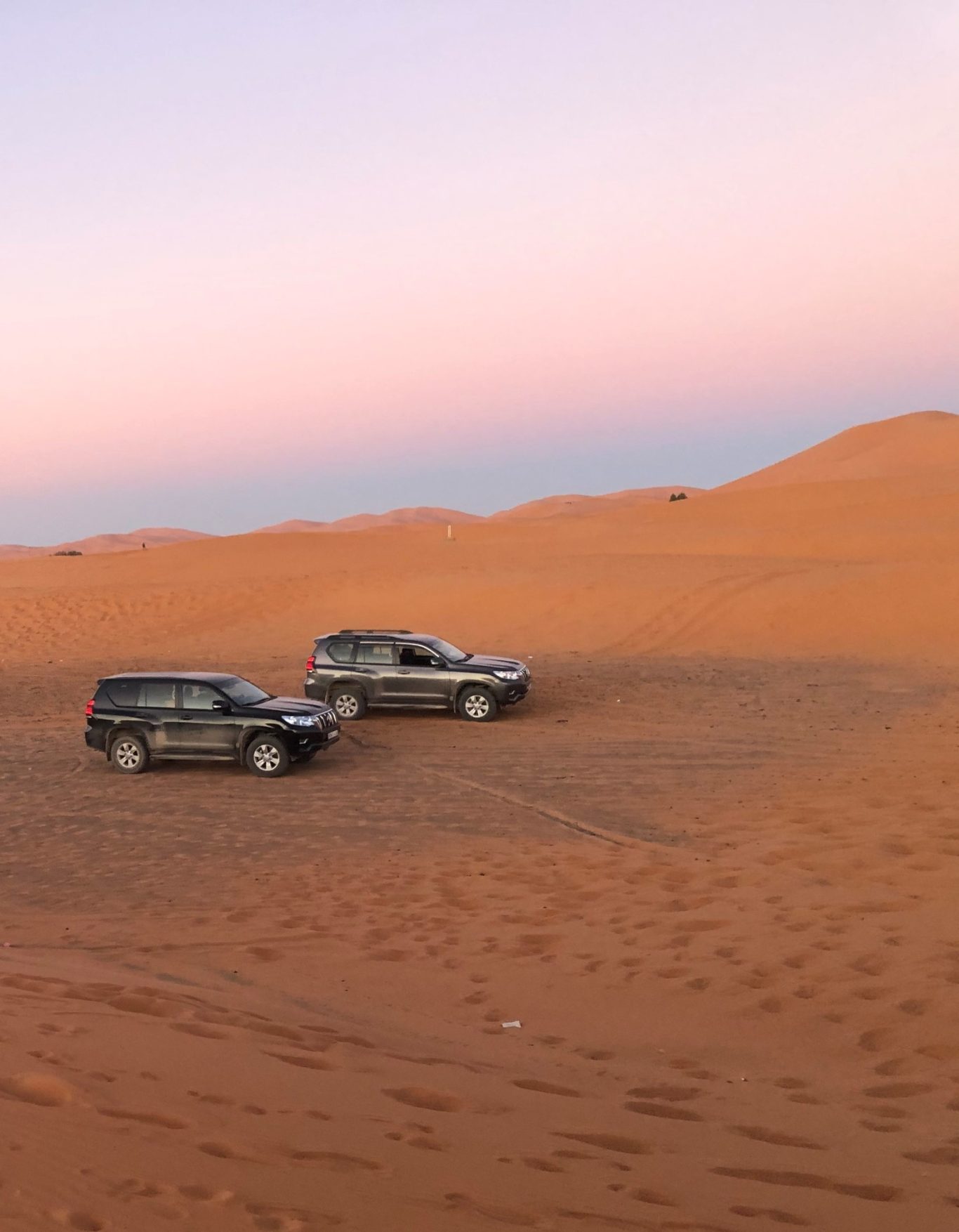 Two four-wheel drive vehicles on sandy dunes at sunset with a pastel sky.