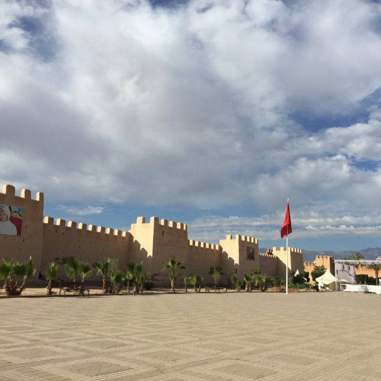 A fortified wall with towers and a red flag, under a cloudy sky.
