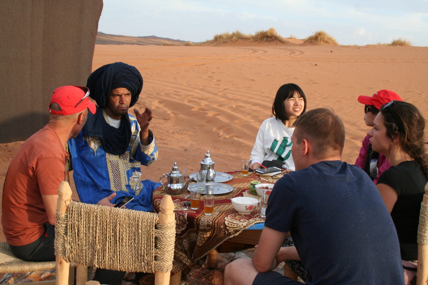 A group of people enjoying tea with a local guide in a desert setting.