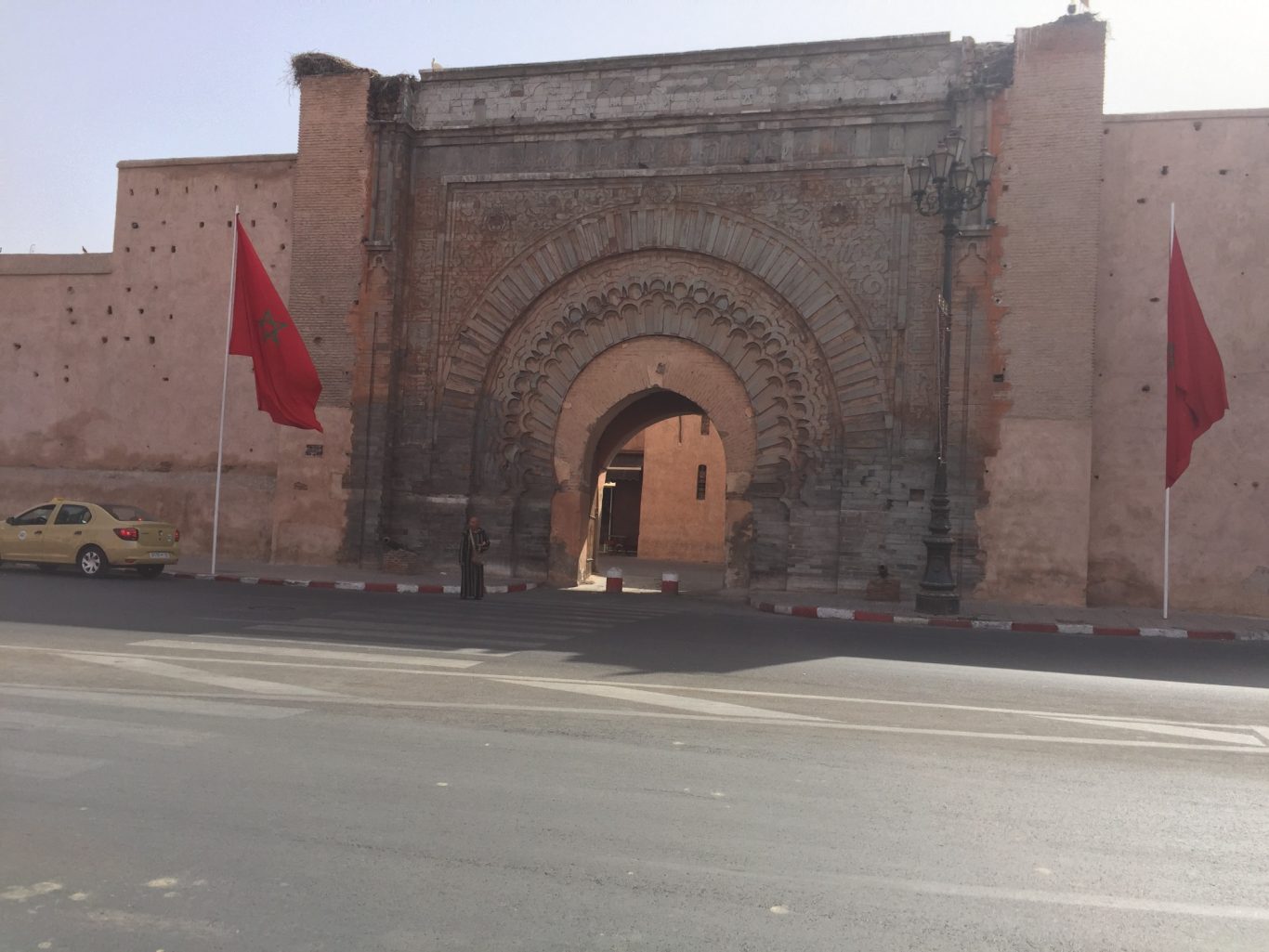 Historic stone gateway with ornate arch, flanked by red flags and a desert backdrop.
