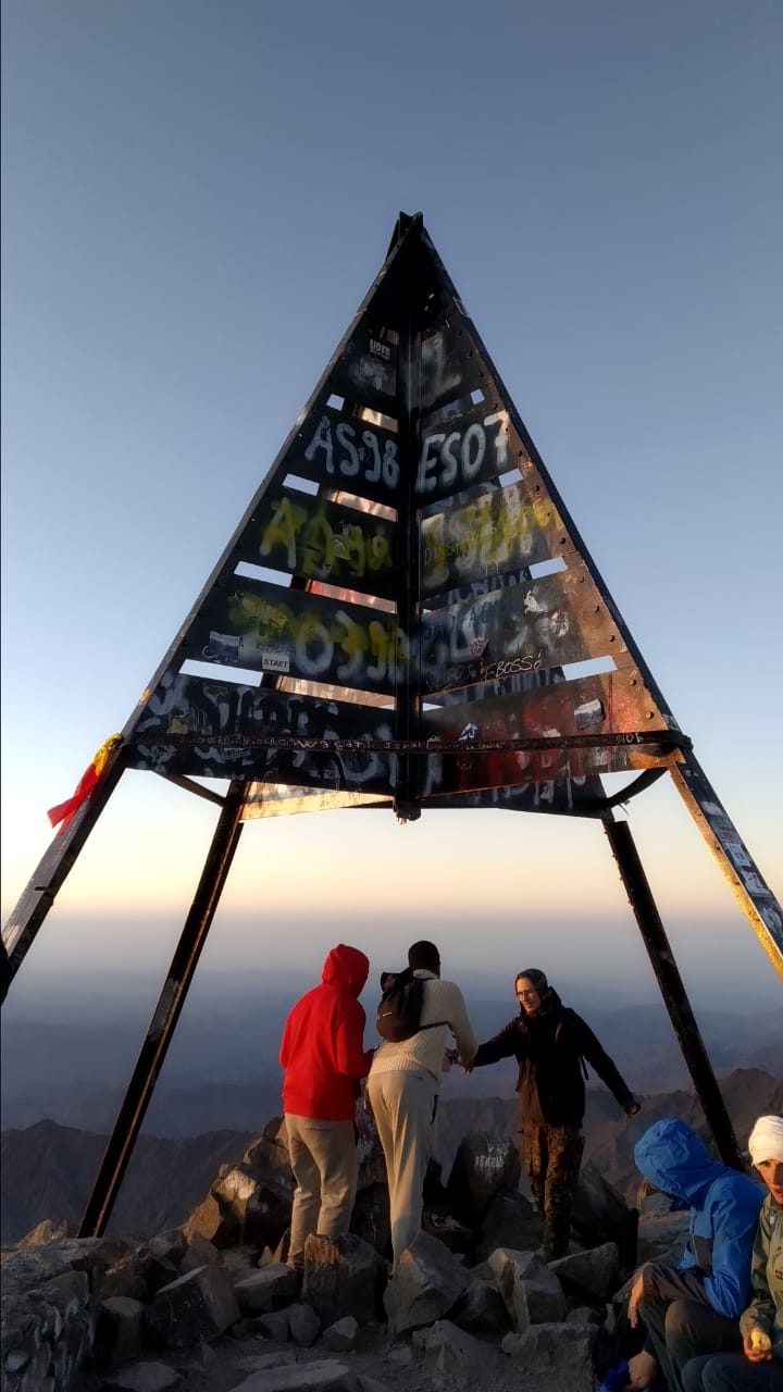 Group of people at the summit of a mountain beneath a large triangular structure.