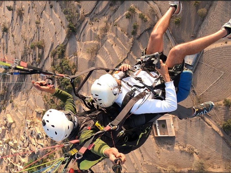 Two people paragliding from a high point with scenic terrain below.