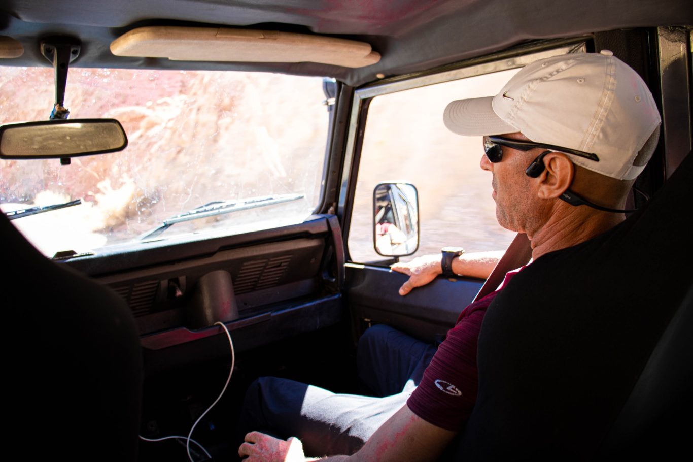 A man wearing a cap and sunglasses sits in a vehicle, looking out the window.