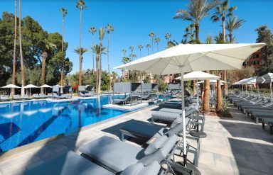 Resort pool area with sun loungers and palm trees under a clear blue sky.