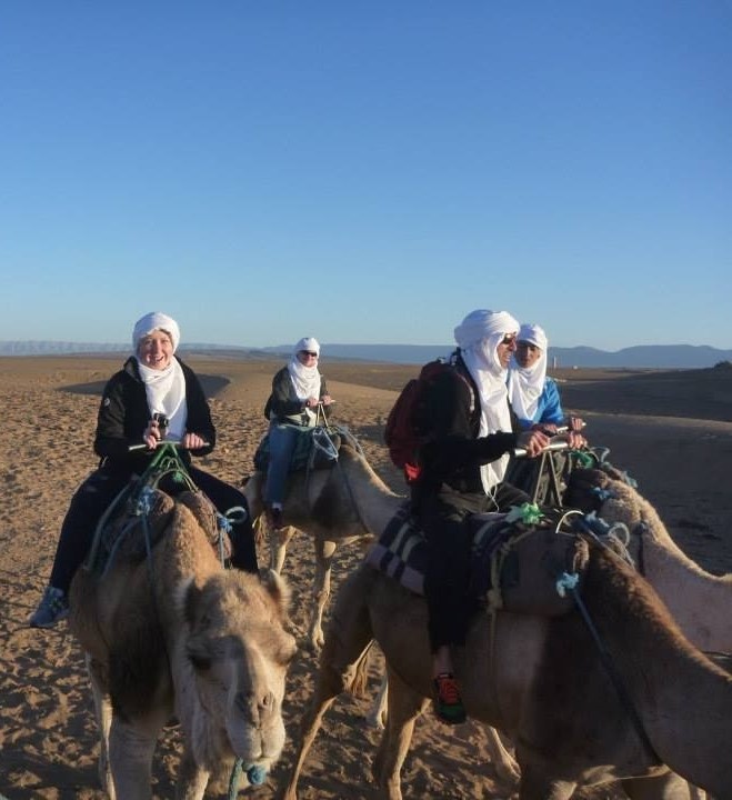Four people riding camels across a sandy desert landscape.
