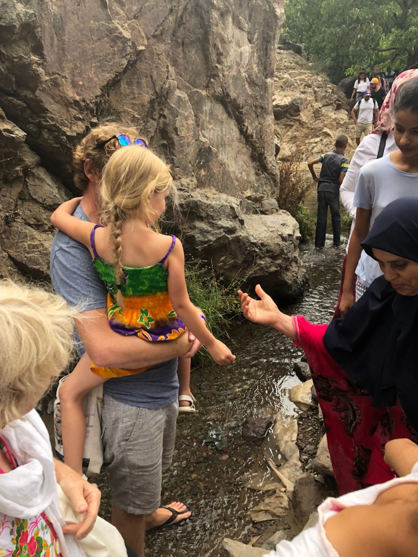 A child in colourful clothing is carried, as adults converse by a stream near rocks.