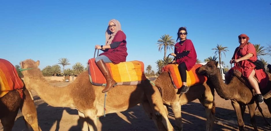 Three people in traditional attire riding camels through a desert landscape.