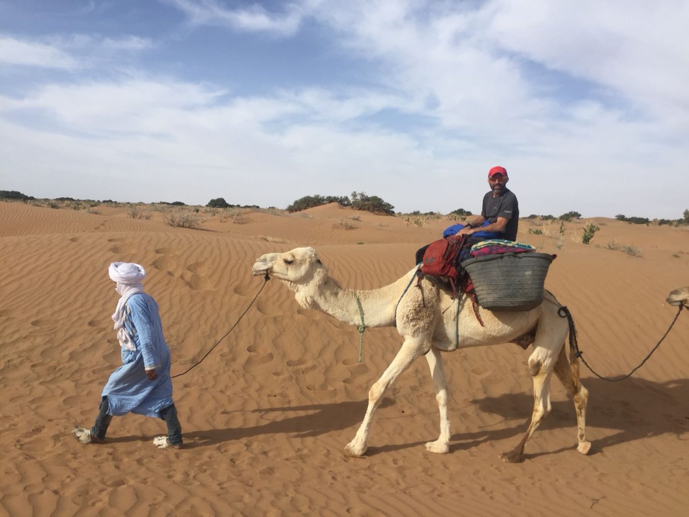A person walks alongside a camel with a rider in a sandy desert landscape.