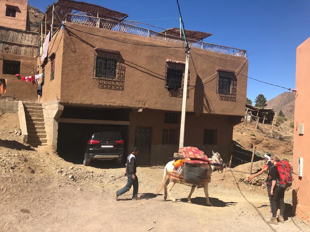 A car parked beside a house, as a person leads a donkey loaded with goods on a dirt path.