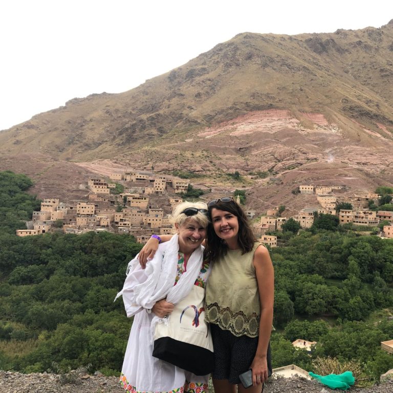 Two women pose together against a mountainous landscape with traditional village homes.
