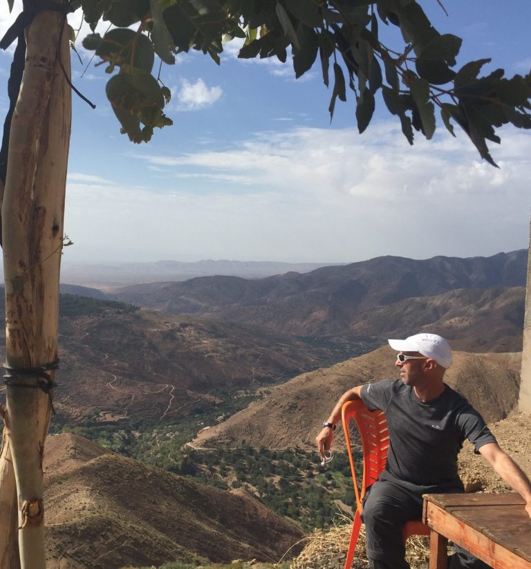 A man sitting on a ledge with a mountain landscape and cloudy sky in the background.