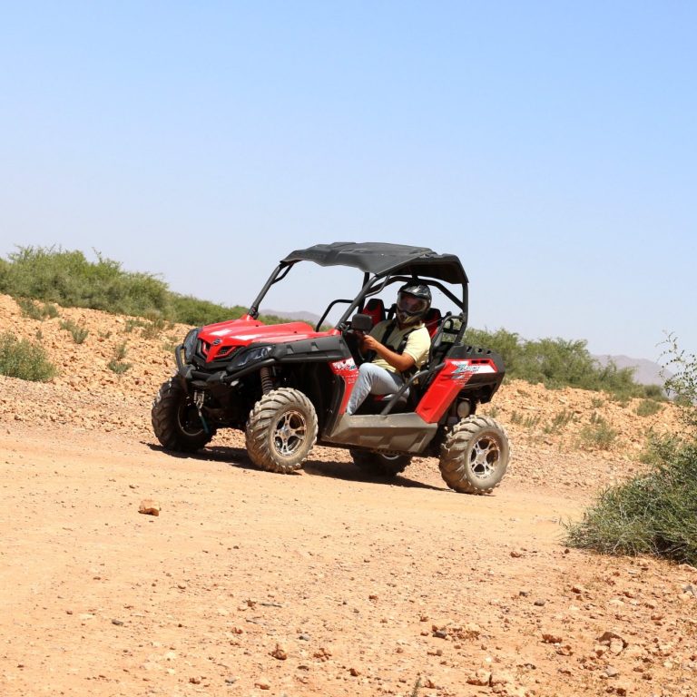 A red off-road vehicle driving on a dirt trail in a desert landscape.