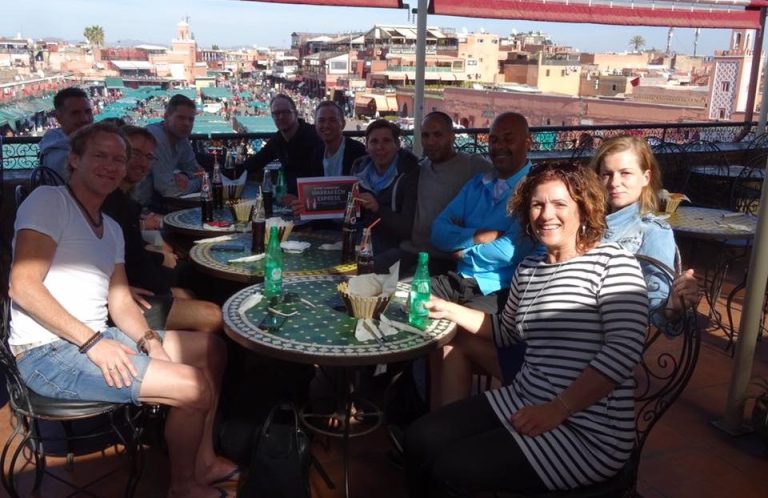 Group of people seated around tables on a rooftop, with a city skyline in the background.