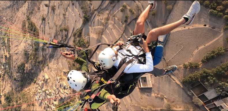Two people tandem paragliding over a landscape, both wearing helmets and harnesses.