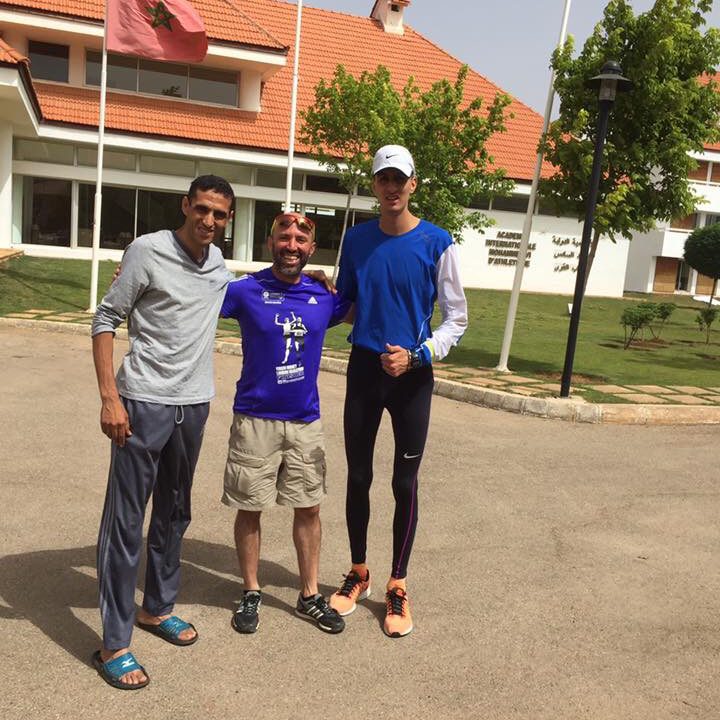 Three men pose together outdoors in front of a building with flags.