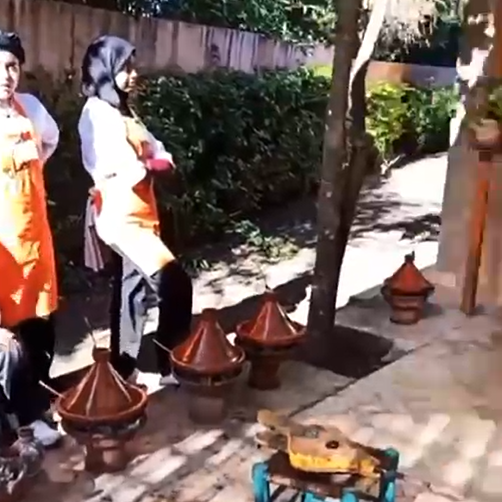 Two people in aprons stand beside traditional clay cooking pots outdoors.