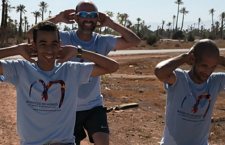 Three men in blue t-shirts playfully covering their ears, smiling outdoors.