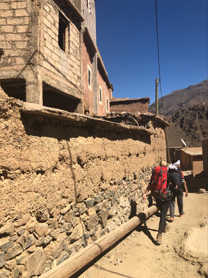 Two hikers walking along a dirt path beside a stone wall in a mountainous area.