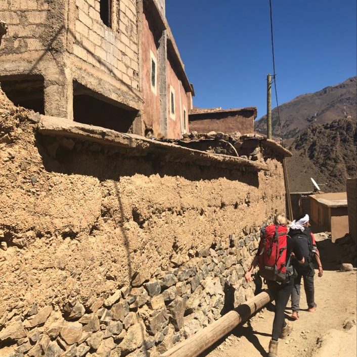 A person walks along a clay wall with a backpack, surrounded by rustic buildings.