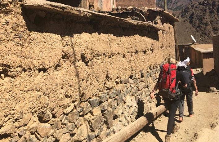 Two hikers walking along a dusty path next to an earthen wall in a rural setting.