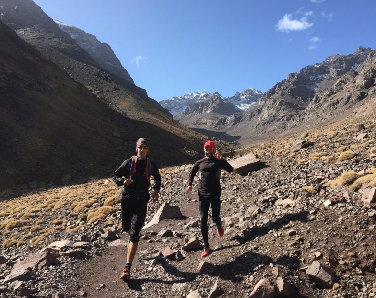 Two runners navigating a rocky mountain trail under a clear blue sky.